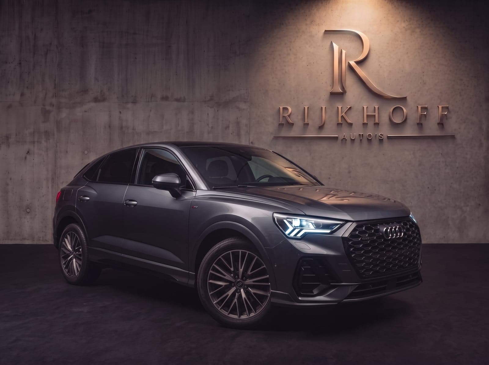 Audi Q3 Sportback in een donkere studio, met het verlichte logo van Rijkhoff Auto's op de betonnen wand
